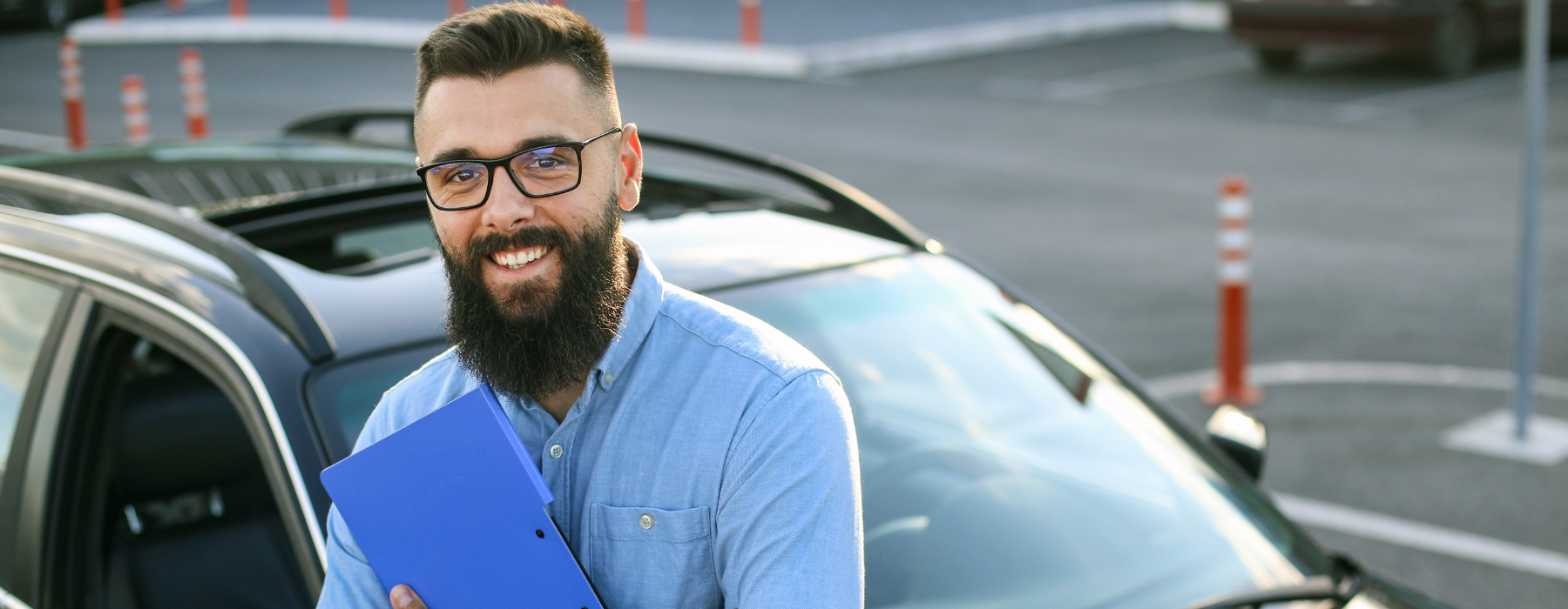 Driving instructor standing by a car holding a clipboard and smiling in a training car park