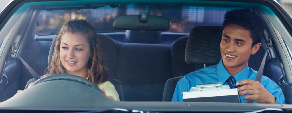 Driving instructor sitting beside a learner driver in a car holding a clipboard during a lesson.