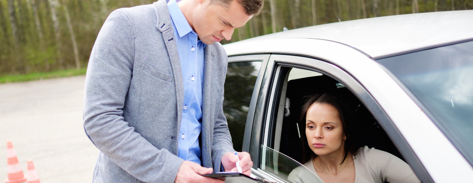 Driving instructor standing beside a dual control car filling out paperwork while a learner driver sits inside during a lesson.